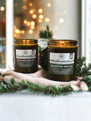 Two lit scented candles in amber glass jars with white labels, one labeled "Yuletide Medley™ - 16oz Twin Wick - By Begonia & Bench®" and the other "Mountain Spruce," are placed on a white surface adorned with evergreen branches. Warm, twinkling lights in the background create a cozy ambiance perfect for fall.