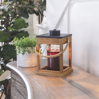 A Wood & Rope Candle Warmer featuring a black top and a sturdy rope handle adorns a wooden surface, exemplifying modern farmhouse decor. A dark red candle is visible inside. To the left, there's a white pot with a small plant. Larger green leaves in the background hint at either garden or indoor plants.