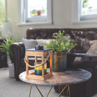 A modern living room showcases a dark brown leather couch adorned with white pillows, exuding a modern farmhouse aesthetic. In the foreground, a round marble coffee table is decorated with the Wood & Rope Candle Warmer and a potted green plant. Soft light streams through large windows, enhancing the calm and cozy atmosphere.