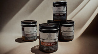 A group of four scented candles in glass jars with black lids are arranged on a brown surface. The labels read "Teakwood Pine," "Smoky Pine," "Desert Spice," and "Rejoy." Each label includes a circular logo and a website link. Shadows cast by the candles create a patterned light effect.