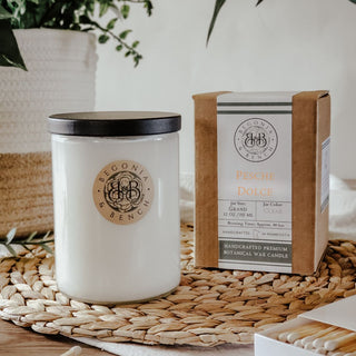 A white candle in a glass jar with a black lid rests on a woven mat. The label reads "Pesche Dolce™ - 12oz Finely Crafted Scented Candle By Begonia & Bench®," suggesting sweet peach aromas. Next to it, there's a brown box labeled with the same inscription, indicating it is a handcrafted premium botanical wax candle. Part of a plant and a matchbox are partially visible.