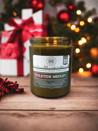 A green scented candle named "Yuletide Medley™ - 16oz Twin Wick - By Begonia & Bench®" in a glass jar sits on a wooden surface. Behind it, a lit Christmas tree with ornaments and red and white wrapped gifts are blurred in the background. A small red ribbon decoration lies to the left of the candle.
