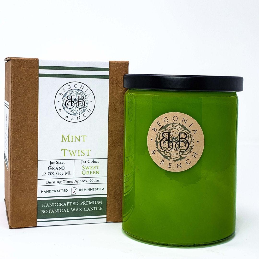 A green candle jar labeled "Mint Twist™" with a "Begonia & Bench®" logo is placed in front of its brown and white packaging box. The packaging details the scented candle's specifications: size (12 oz), color (sweet green), burn time (approximately 90 hours), and origin (Minnesota).