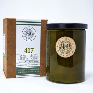 A vintage green glass candle jar with a black lid stands beside a brown cardboard box. Both the jar and box feature the brand "Begonia & Bench." The box details include "417™ - 12oz Finely Crafted Scented Candle By Begonia & Bench®," burning time (80 hrs), and mentions the scented candle is handcrafted in Minnesota.
