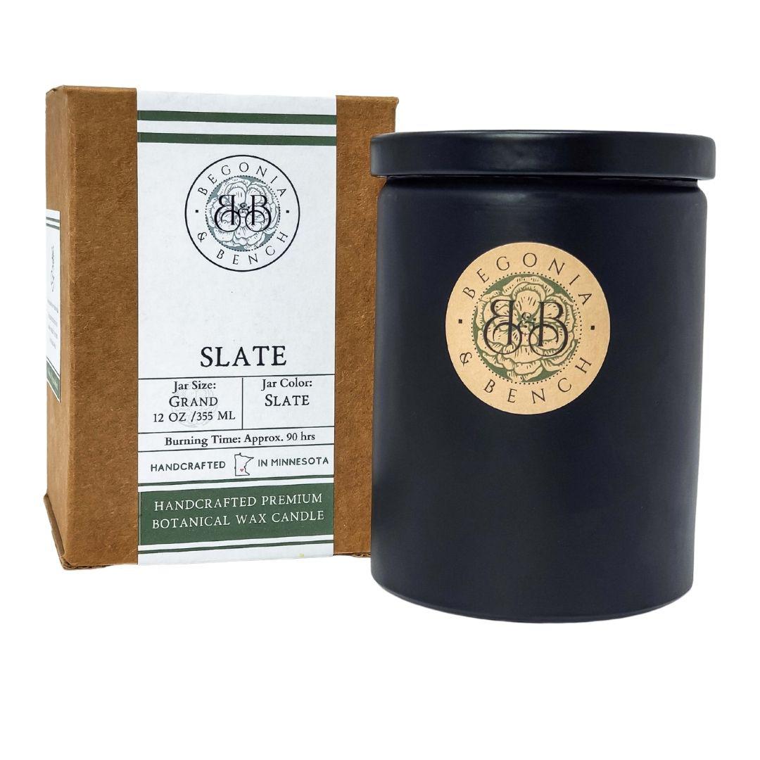 A black candle jar with a beige label featuring "Begonia & Bench" and their logo. The brown box packaging beside it reads "Slate™ - 12oz Finely Crafted Scented Candle By Begonia & Bench®," specifying the grand size of 12 oz / 355 ml and jar color as slate. This handcrafted scented candle offers a burn time of approximately 90 hours, made from premium botanical wax.