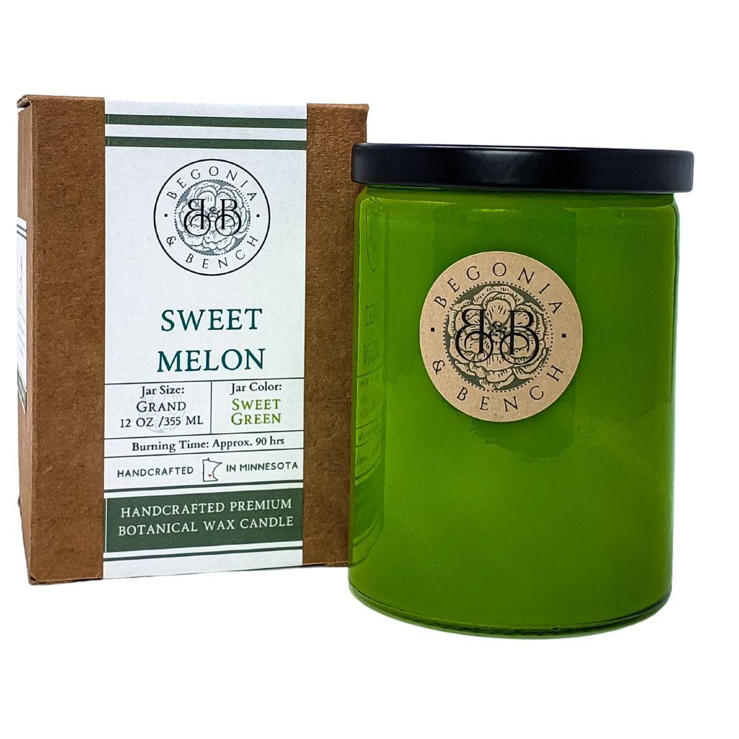 A green Sweet Melon™ - 12oz Finely Crafted Scented Candle by Begonia & Bench®, featuring a black lid and labels of "Begonia & Bench®" and "Sweet Melon," stands next to a brown box of the same brand. The box details this fall release: grand jar size, 12 oz/355 ml, sweet melon scent, green color, handcrafted premium botanical wax, with an approximately 80-hour burn time.