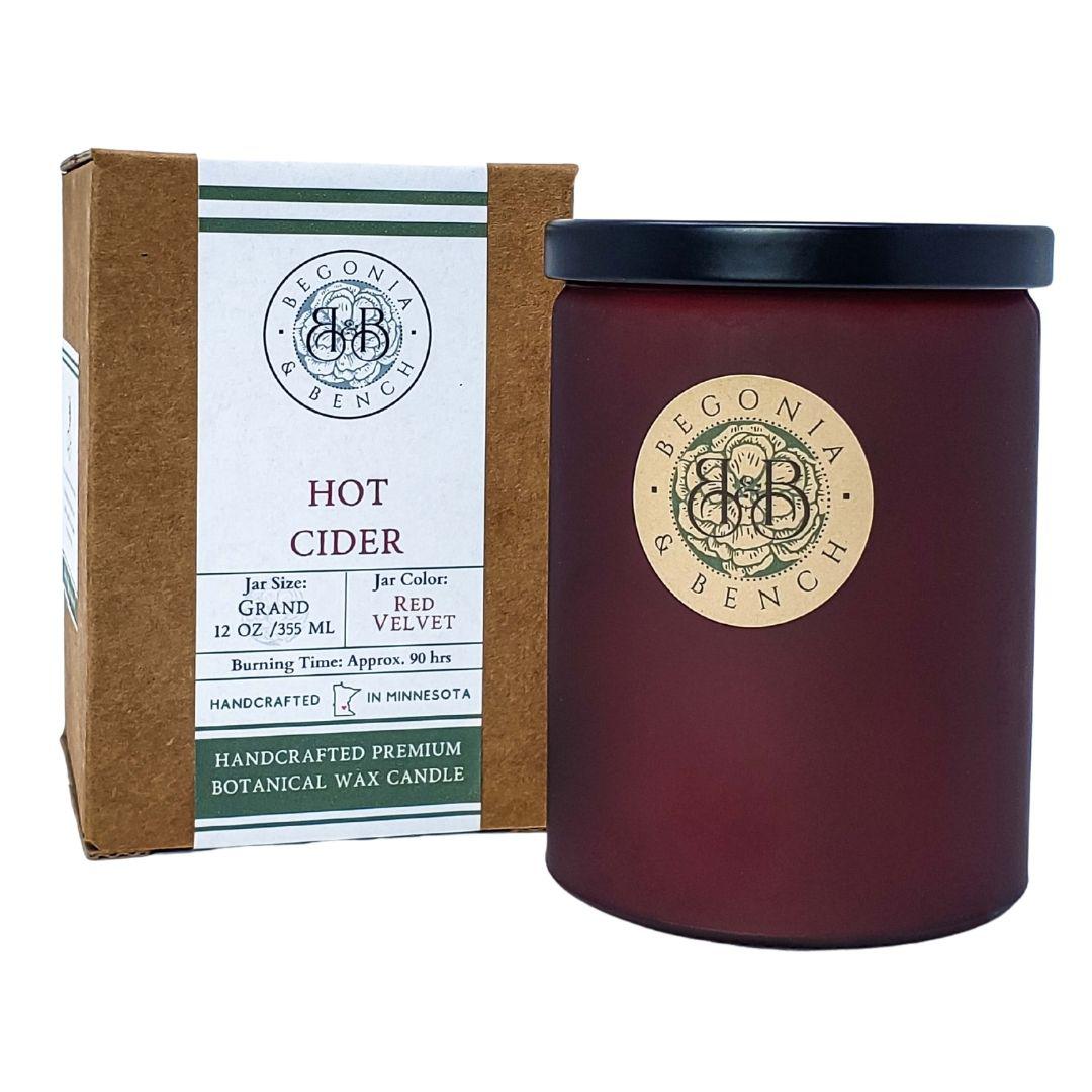 A Hot Cider™ - 12oz Finely Crafted Scented Candle by Begonia & Bench® is displayed, exuding a hot cider aroma. The deep red candle jar with a black lid features an emblem on the front. Next to it sits the brown packaging box, adorned with green accents and product details like size, fragrance, and burning time.