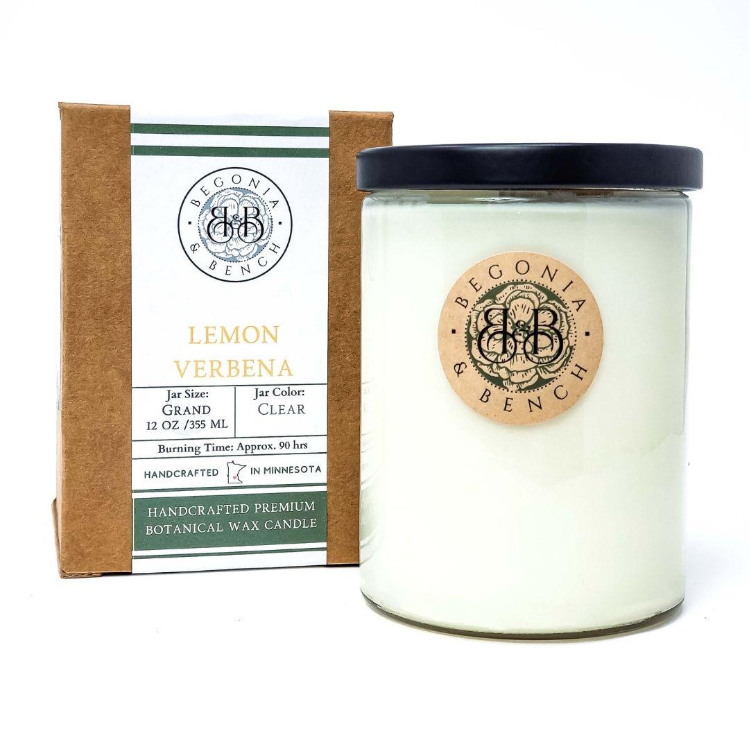 A finely crafted lemon verbena scented candle from Begonia & Bench® rests in a clear jar with a black lid beside its brown and green packaging box. The box reads "Begonia Bench, Lemon Verbena, Grand Size 12 oz, 355 ml, Burning Time Approx. 90 hrs" and "Handcrafted in Minnesota." Both the jar and box feature a wooden emblem displaying the brand logo.