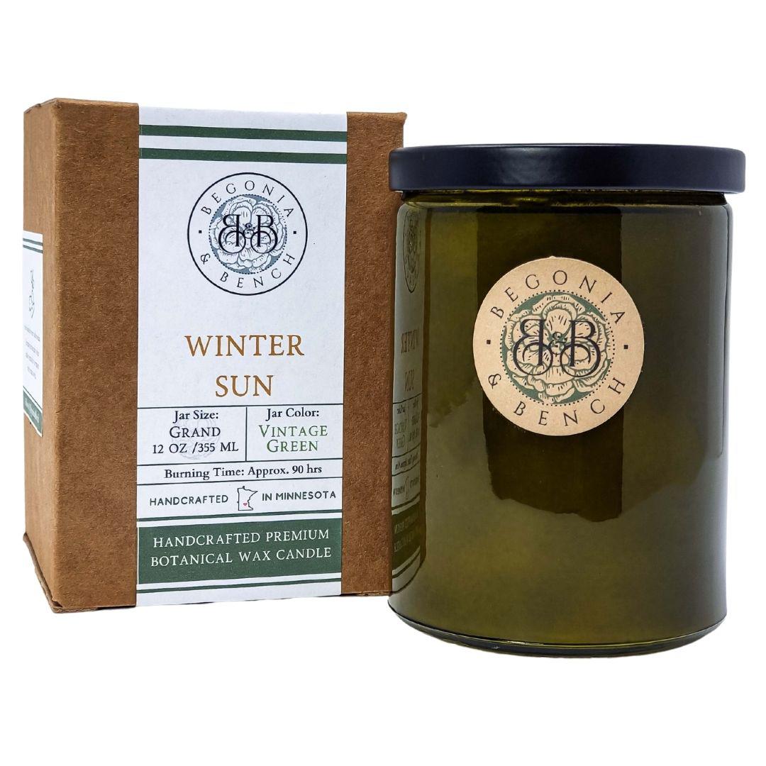 A brown box with a white label reading "Begonia & Bench," "Winter Sun," and detailing the 12 oz (355ml) size and 90-hour burn time is displayed alongside a green glass fall candle jar. The scented candle jar, topped with a black lid and adorned with a matching round label, is handcrafted in Minnesota and known as Winter Sun™ - a 12oz finely crafted scented candle by Begonia & Bench®.