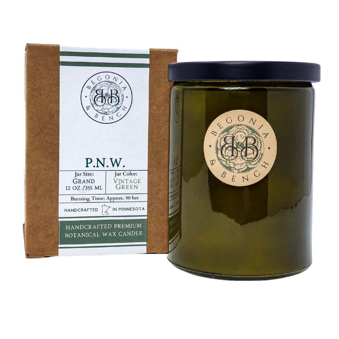 Image shows a P.N.W. - 12oz Finely Crafted Scented Candle by Begonia & Bench®. This fall candle, scented with natural botanicals, comes in a vintage green 12 oz glass jar with a black lid. The label on the brown box provides details about the jar size, color, and an approximate burning time of 90 hours. Handcrafted in Minnesota.
