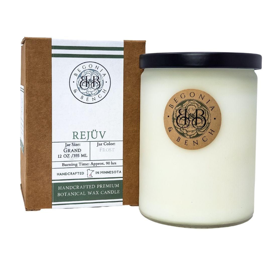 A white scented candle in a frosted glass jar with a black lid sits next to a brown and white box. Both the jar and the box feature labels for "Begonia & Bench®" and their product "Rejüv™ - 12oz Finely Crafted Scented Candle." The label indicates the jar size is 12 oz, with an approximate burning time of 90 hours.