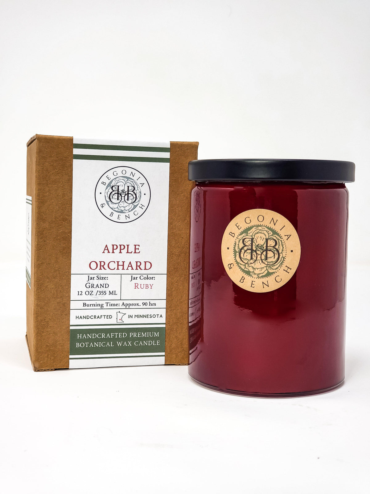 A cylindrical ruby-colored *scented candle* in a glass jar, branded as Begonia & Bench®, stands next to its packaging. The label on the jar and box reads "Apple Orchard." The box details include jar size (12 oz/355 ml), estimated burning time (90 hrs), and that it is handcrafted in Minnesota.