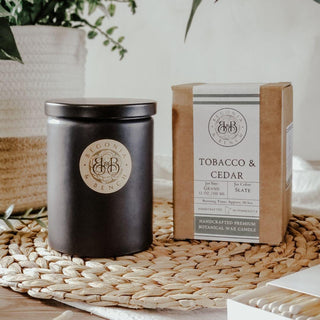 A black candle jar labeled "Begonia & Bench" sits on a woven mat, exuding fall candle vibes. To its right, a tan box with the same brand name reads "Tobacco & Cedar™ - 12oz Finely Crafted Scented Candle By Begonia & Bench®" and details the scented candle's aroma and size. In the background, a potted plant and a white object rest on a light-colored surface.