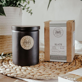 A black jar candle with a beige label displaying "Begonia & Bench" stands next to its packaging. The box, labeled "Slate™ - 12oz Finely Crafted Scented Candle By Begonia & Bench®," describes it as a handcrafted premium botanical wax fall candle, 12 oz/345 ml. Both items are on a woven mat with greenery nearby.