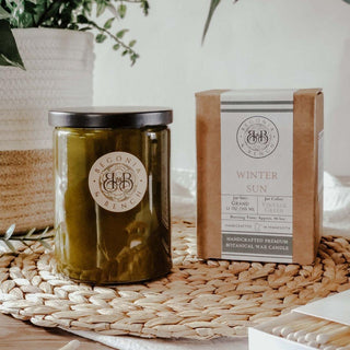 A finely crafted 12oz scented candle labeled "Winter Sun™" by Begonia & Bench® sits in its vintage green glass jar next to its matching box on a natural woven placemat. The scene is bright and cozy, with a hint of greenery in the background, evoking a calm and inviting atmosphere perfect for fall.