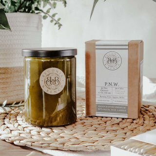 A green glass P.N.W. 12oz finely crafted scented candle by Begonia & Bench®, featuring a black lid, is placed on a woven mat. Beside it is a brown box labeled “Begonia & Bench,” which details the scent as P.N.W. and highlights that this handcrafted candle is made of botanical wax with notes of fir and pine. Greenery is visible in the background.