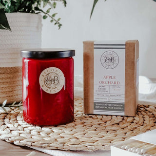 An Apple Orchard - 12oz Finely Crafted Scented Candle By Begonia & Bench® in a red glass jar with a silver lid sits next to its cardboard packaging on a woven placemat. The packaging features the label "Begonia & Bench®," along with product details about the handcrafted, premium botanical wax candle. Greenery and a light background are also visible.