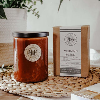 A hand-poured soy scented candle in a glass jar with a black lid is placed on a woven mat. The jar label reads "Begonia & Bench." Next to it is a rectangular kraft paper box labeled "Morning Blend™ - 12oz Finely Crafted Scented Candle By Begonia & Bench®" that describes the scent and details of this cozy fall candle. In the background are greenery and soft furnishings.