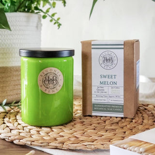 A green jar candle labeled "Begonia Bench" is positioned on a woven mat. Next to it is the candle's box marked "Sweet Melon™," indicating it is a finely crafted botanical wax scented candle, 12 oz, by Begonia & Bench®, with details about scent and craftsmanship. A plant and some matches are in the background.