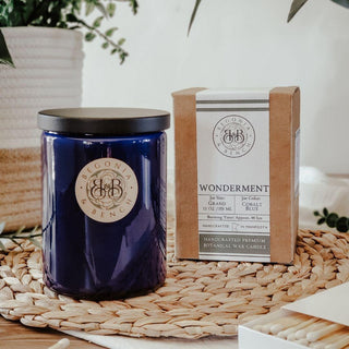 A cobalt blue 12oz finely crafted scented candle by Begonia & Bench®, labeled "Wonderment™", sits in a glass jar with a black lid beside its tan and white packaging box. The box displays the brand names "Begonia & Bench®" and "Wonderment candles" along with detailed product information. Both the candle and its packaging are placed on a woven mat, with some greenery in the background, emphasizing their exotic aromas.