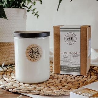 The image displays a white botanical scented candle in a glass jar with a black lid, labeled as "Peppercorn - 12oz Finely Crafted Scented Candle by Begonia & Bench®." Next to the candle is its brown box packaging which provides additional details about the candle's size, weight, and ingredients. Both items are placed on a woven mat with a leafy plant in the background.
