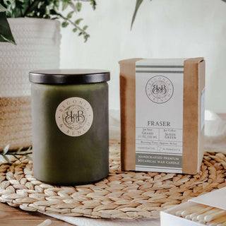 A green fall candle in a glass jar with a black lid sits beside its packaging on a woven mat. Both the jar and the box are labeled "Begonia & Bench®," with the text "Fraser™ - 12oz Finely Crafted Scented Candle" visible on the box. A potted plant and a white scented candle in the background add to the cozy, natural setting.