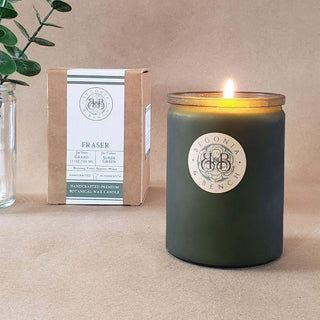 A lit Fraser™ - 12oz finely crafted scented candle by Begonia & Bench® in a dark green jar with a beige label is in the foreground. To the left, a brown cardboard box bears the same brand and product information. A sprig of greenery in a glass jar is partially visible in the top left corner.