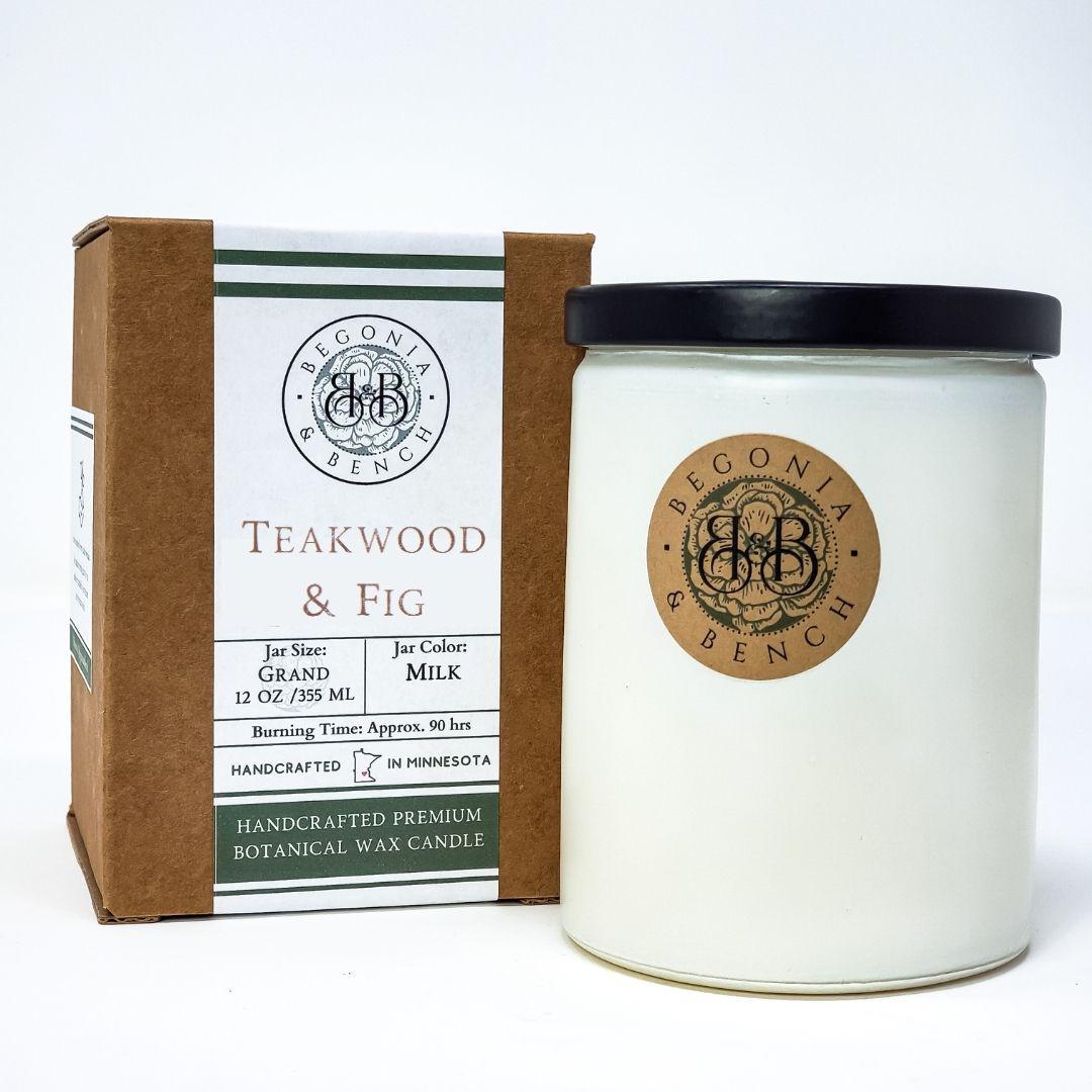A white scented candle in a cylindrical glass jar with a black lid, branded "Begonia & Bench," is labeled "Teakwood & Fig, Jar Size: Grand, 12 oz / 355 ml, Jar Color: Milk, Burning Time: Approx. 90 hrs, Handcrafted in Minnesota." This fall candle is positioned beside its brown cardboard packaging. Officially named "Teakwood & Fig™ - 12oz Finely Crafted Scented Candle By Begonia & Bench®.