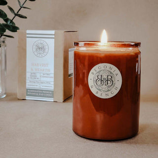 A lit brown jar candle featuring the "Begonia & Bench®" label rests on a beige surface. To the left, there's a cardboard box labeled "Harvest & Hearth™ - 12oz Finely Crafted Scented Candle By Begonia & Bench®," describing it as a handcrafted premium botanical wax fall candle. In the background, green foliage in a glass jar is blurred.