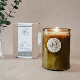 A lit P.N.W. - 12oz Finely Crafted Scented Candle by Begonia & Bench® in a green glass jar sits on a surface next to a cardboard box. The box displays details about the fall candle, including "Grand Fir" and "Vintage Green." A clear glass vase with green leaves is in the background.