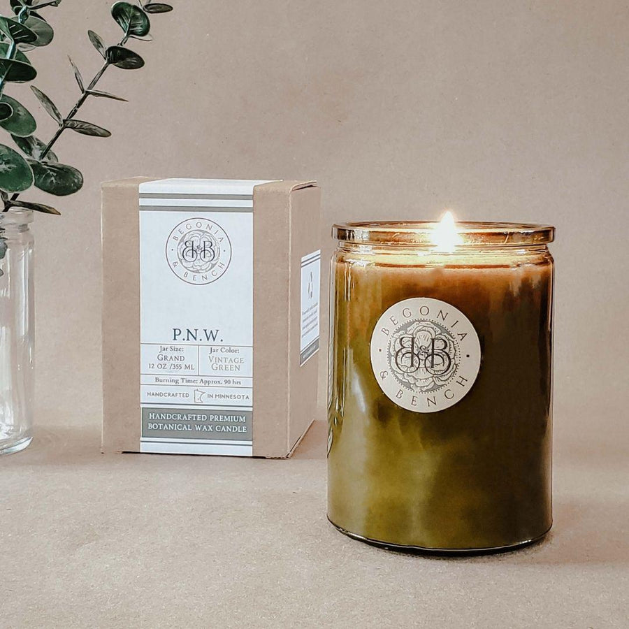 A lit P.N.W. - 12oz Finely Crafted Scented Candle by Begonia & Bench® in a green glass jar sits on a surface next to a cardboard box. The box displays details about the fall candle, including "Grand Fir" and "Vintage Green." A clear glass vase with green leaves is in the background.