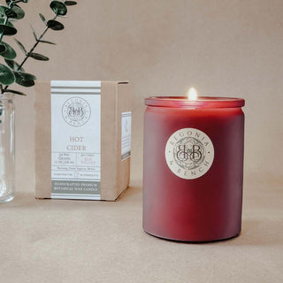 A lit, burgundy-colored fall candle in a glass jar bearing the "Begonia & Bench®" label rests on a beige surface. To the left, there is a plant in a transparent glass vase and a cardboard box with the same branding and text reading "Hot Cider™ - 12oz Finely Crafted Scented Candle," indicating the candle's inviting aroma. The background is neutral.