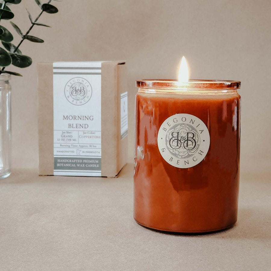 A finely crafted 12oz scented candle from Begonia & Bench®, labeled "Morning Blend™," is lit and placed on a light brown surface in a glass container. Behind it, a box bearing the same label is visible. The background features a clear vase with green foliage on the left, creating an ideal ambiance for fall.