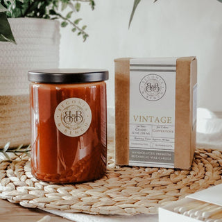 A brown candle jar with a black lid sits next to a box labeled "Vintage™ - 12oz Finely Crafted Scented Candle By Begonia & Bench®" on a woven placemat. The fall candle features the Begonia & Bench® logo and the box states it's a handcrafted premium botanical wax scented candle in Coppertone scent. A plant and woven basket are in the background.