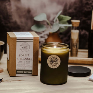 A green glass candle from Begonia & Bench®, labeled "Forest & Flannel™ - 12oz Finely Crafted Scented Candle," burns with a warm flame on a wooden table. Next to it is a brown box describing the fall candle's inviting scent. The background features a blurred image of plants and wooden objects, creating a cozy atmosphere.