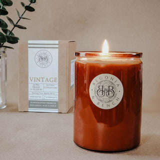 A lit brown jar candle labeled "Vintage™ - 12oz Finely Crafted Scented Candle By Begonia & Bench®" is placed on a beige surface. Behind it, there is a brown box with a white label that provides details about the scented candle's make and aroma. A green leafy plant in a glass vase is partially visible in the background.