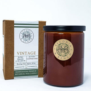 A brown glass jar candle labeled "Vintage™ - 12oz Finely Crafted Scented Candle By Begonia & Bench®" is placed next to its brown cardboard box packaging. The box label reads "Vintage" and provides details such as jar size (12 oz/355 ml), jar color (Coppertone), and burning time (approx. 90 hrs). Handcrafted in Minnesota, this scented fall candle adds a cozy touch to any space.