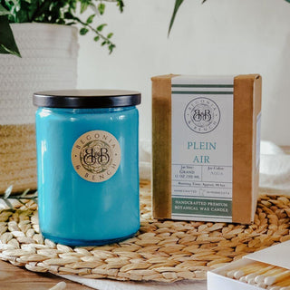 A blue scented candle in a glass jar with a lid, labeled "Plein Air™ - 12oz Finely Crafted Scented Candle By Begonia & Bench®," sits on a woven mat. Next to it is a cardboard box with "Plein Air," indicating the scent and details of the handcrafted premium botanical wax candle. Background includes houseplants and a wooden surface.