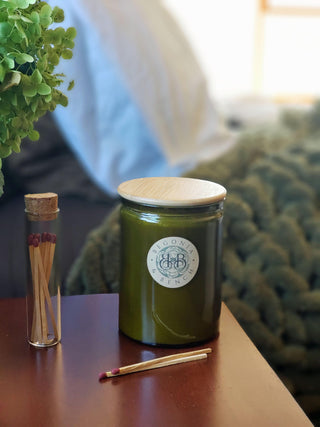 A fall candle with a green hue and a bamboo lid labeled "Begonia Esencia" sits on a wooden table next to a glass jar filled with matches. One match lies separately on the table. In the background, a green textured blanket and plant leaves are partially visible, hinting at a cozy, serene setting.