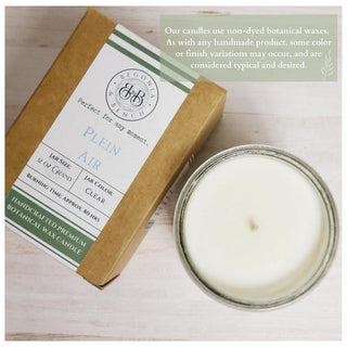 Top view of a Citrus & Clove™ scented candle in a brown cardboard box beside the candle in a glass jar. The label on the box reads "Plein Air" and "Handcrafted Premium Botanical Wax Candle." A circular note indicates variations due to handmade nature. Wooden surface background. Suitable as a fall candle by Begonia & Bench®.