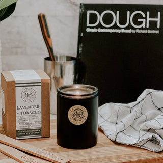A black candle jar labeled "Lavender + Tobacco™ - Signature Aroma by Waybread" sits on a wooden surface next to its box. There's a cookbook titled "DOUGH" in the background. A kitchen towel is draped on the right, and a metal utensil holder with wooden tools inside is partially visible—the perfect ambiance for a fall scented candle.