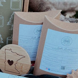The image shows two eco-friendly, brown paper packages with white labels from Blooma Box. One is marked "12 oz Grand Lake" and the other "Minnesota Engraved Bamboo Lid." A round bamboo coaster displaying a silhouette of Minnesota with a heart in the center is placed in the foreground alongside a fall candle.