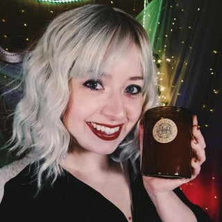 A smiling person with wavy, shoulder-length blonde hair and dark red lipstick holds up a Forest & Flannel™ - 12oz Finely Crafted Scented Candle by Begonia & Bench® in a dark glass jar. The background is decorated with colorful fabric, string lights, and fairy lights, creating a festive atmosphere. The person is wearing a dark top and a nose ring.