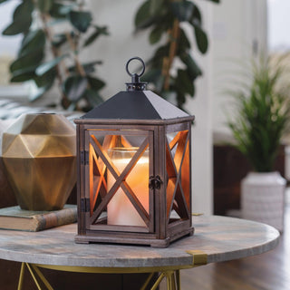 A rustic lantern with a glowing candle inside sits on a round marble table. The lantern has crosshatched glass panes and a black metal roof with a loop handle. In the background, there is greenery, a geometric gold vase, and a white textured plant pot, creating a cozy atmosphere.