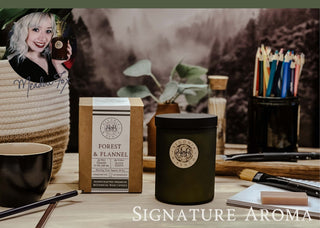 A cozy scene features a "Forest & Flannel™ - 12oz Finely Crafted Scented Candle By Begonia & Bench®" in a dark green glass container, complemented by a matching box and various desk items such as a potted plant, colored pencils, and a coffee cup. An inset photo in the top-left corner shows a smiling woman holding the scented candle. The text reads "Signature Aroma".