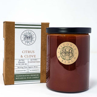 A 12 oz Citrus & Clove™ scented candle jar with dark brown wax and a black lid labeled "Begonia & Bench®" stands next to its brown packaging box. The box provides information on the jar size, color, burn time (90 hrs), and its handcrafted origin in Minnesota.