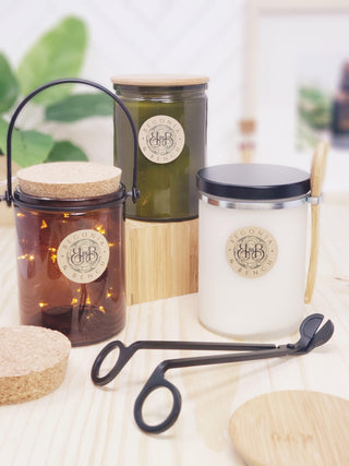 Three scented candles are displayed, each in jars of different colors: amber, green, and white. The jars are labeled "Geconia & Bench" and have cork lids. Additional candle accessories, such as a wick trimmer and a wick snuffer, are placed in front of the candles on a wooden surface along with a Bamboo Spoon & Clip.